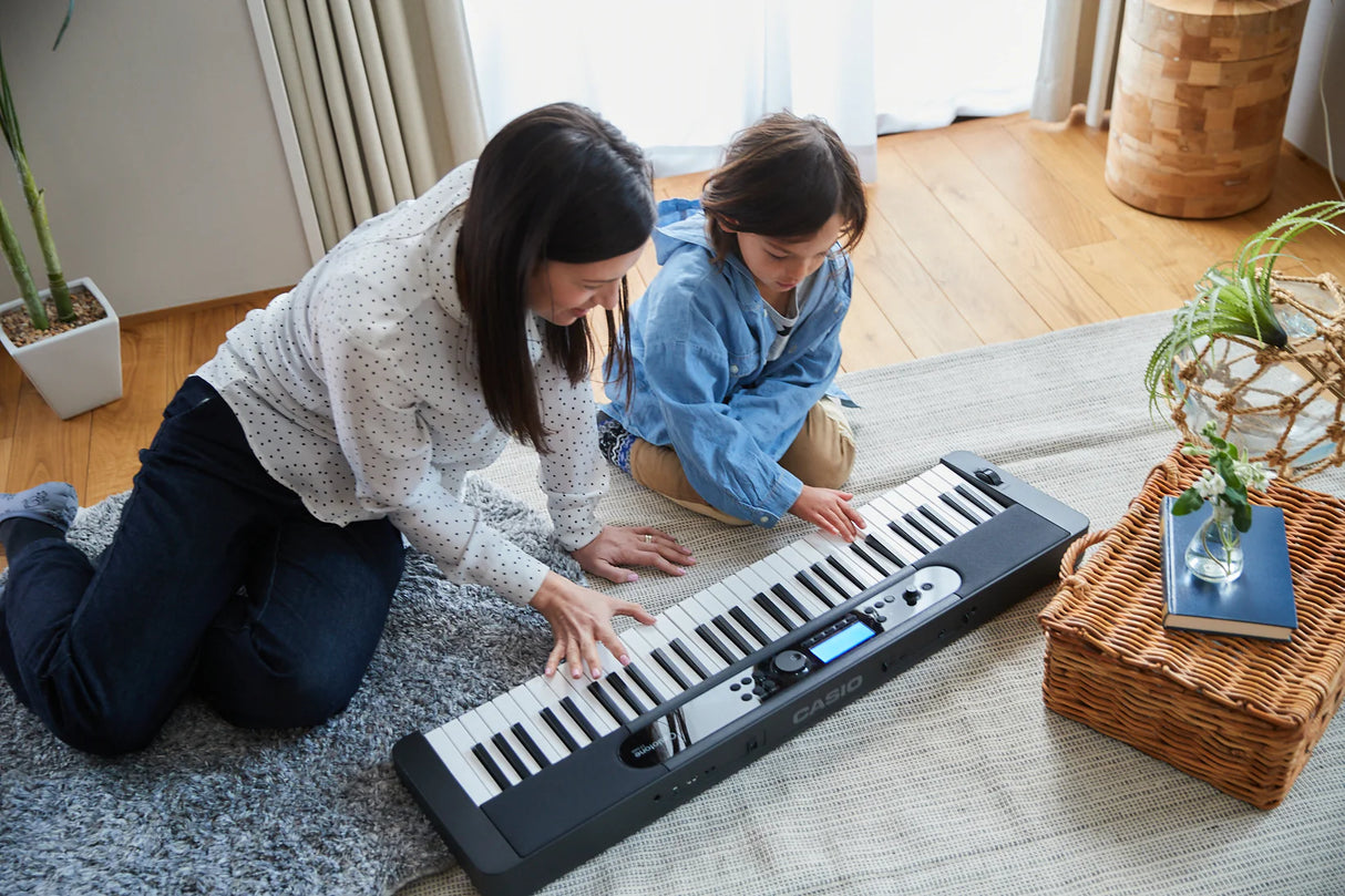 Two people playing a keyboard on a carpeted floor in a room with plants and a basket.