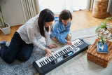 Two people playing a keyboard on a carpeted floor in a room with plants and a basket.